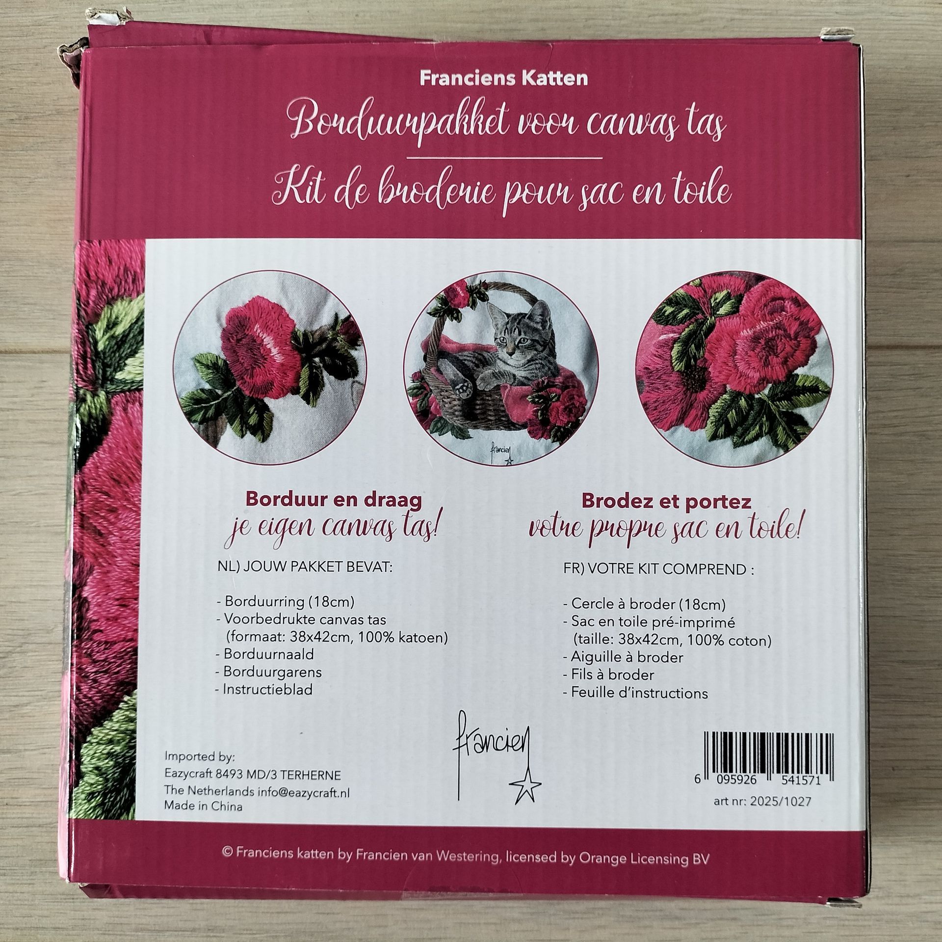 Een borduurpakket in een doos met een kat in een mandje, omringd door rozen, op een draagtas. Roze en wit.