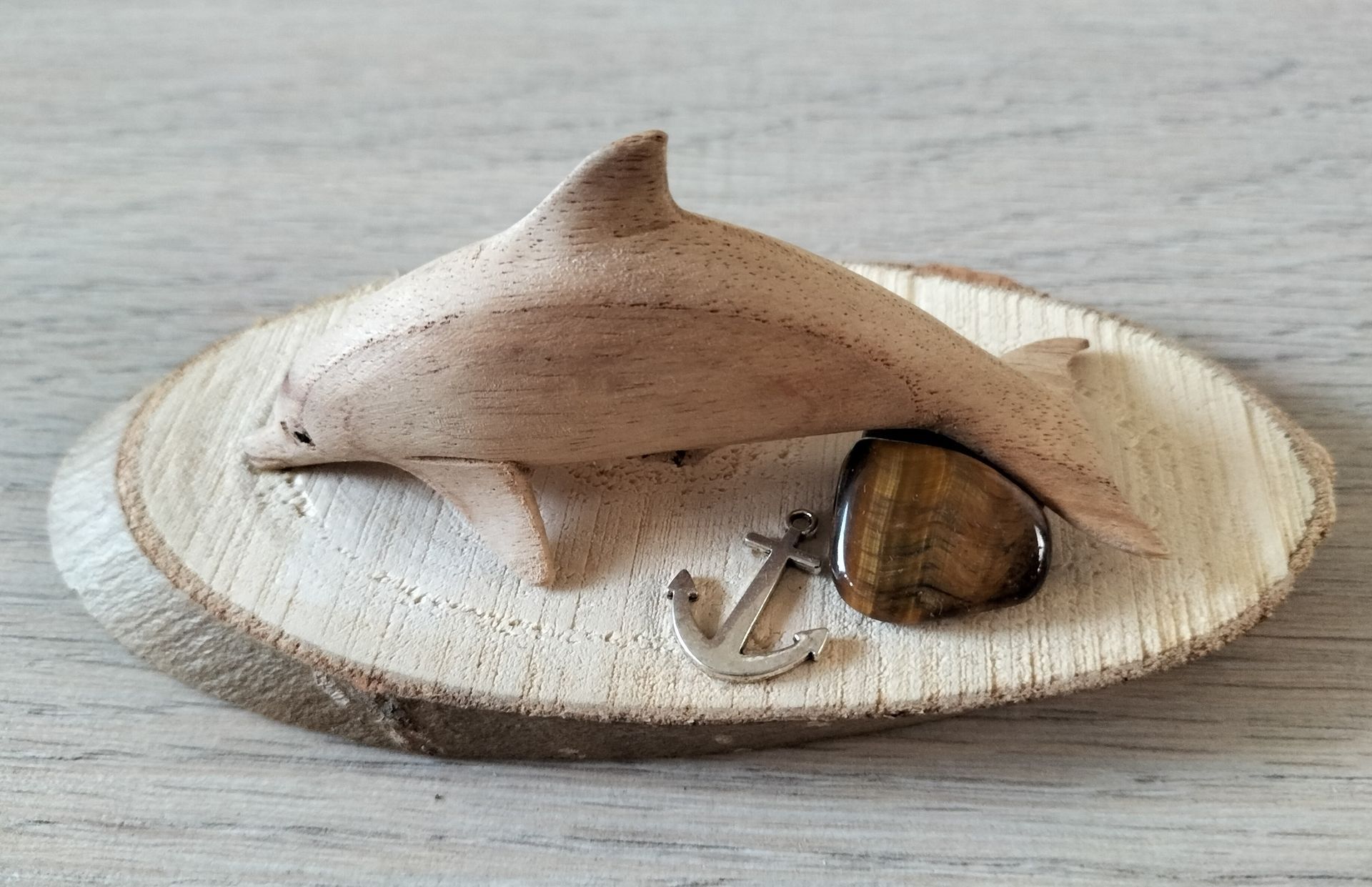 Een houten dolfijnsculptuur op een houten schijf als sokkel, met een tijgeroogsteen en een klein anker.