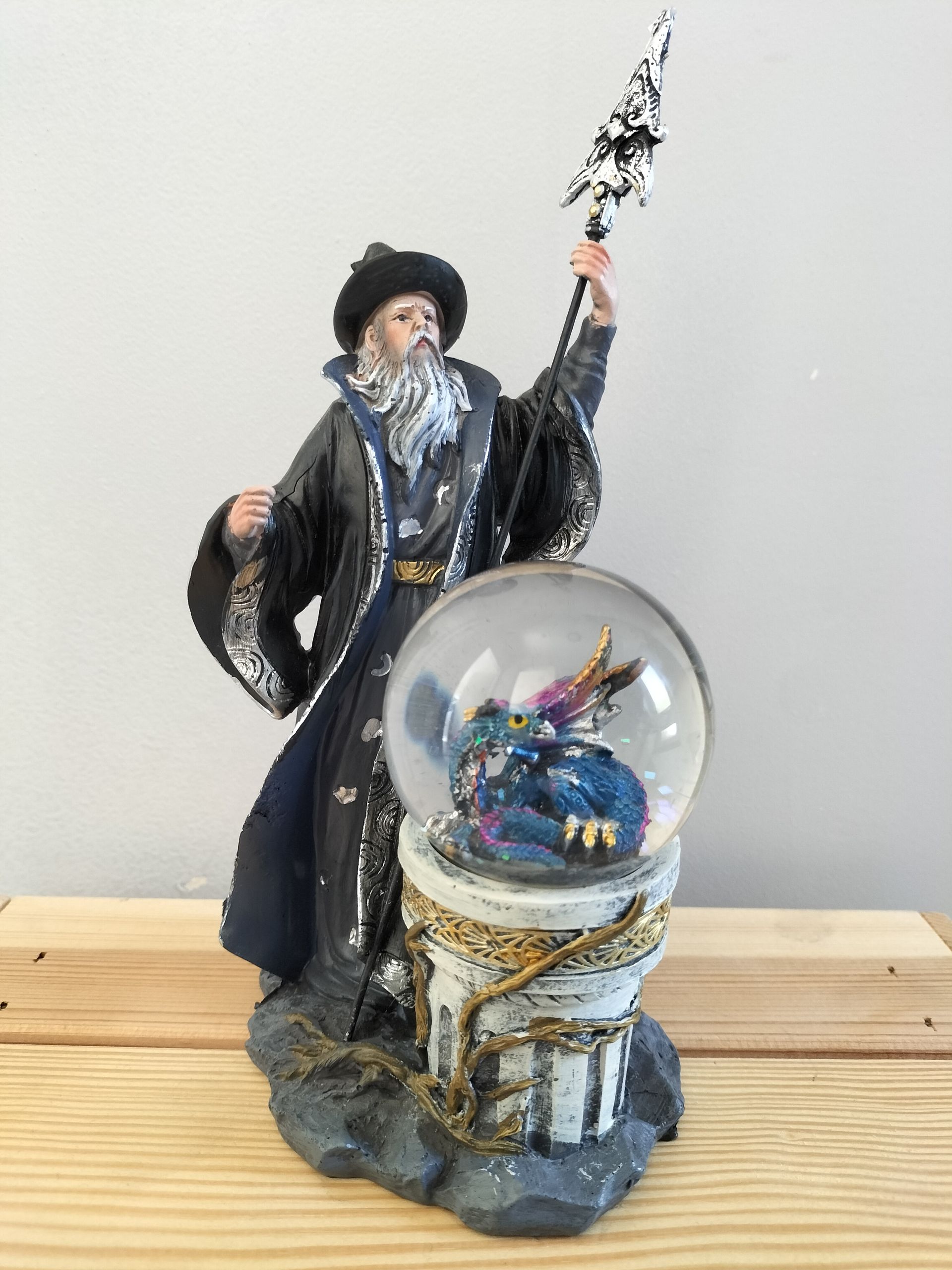 Beeld van een tovenaar in zwarte mantel met een staf, naast een sneeuwbol met een draak op een pilaar.