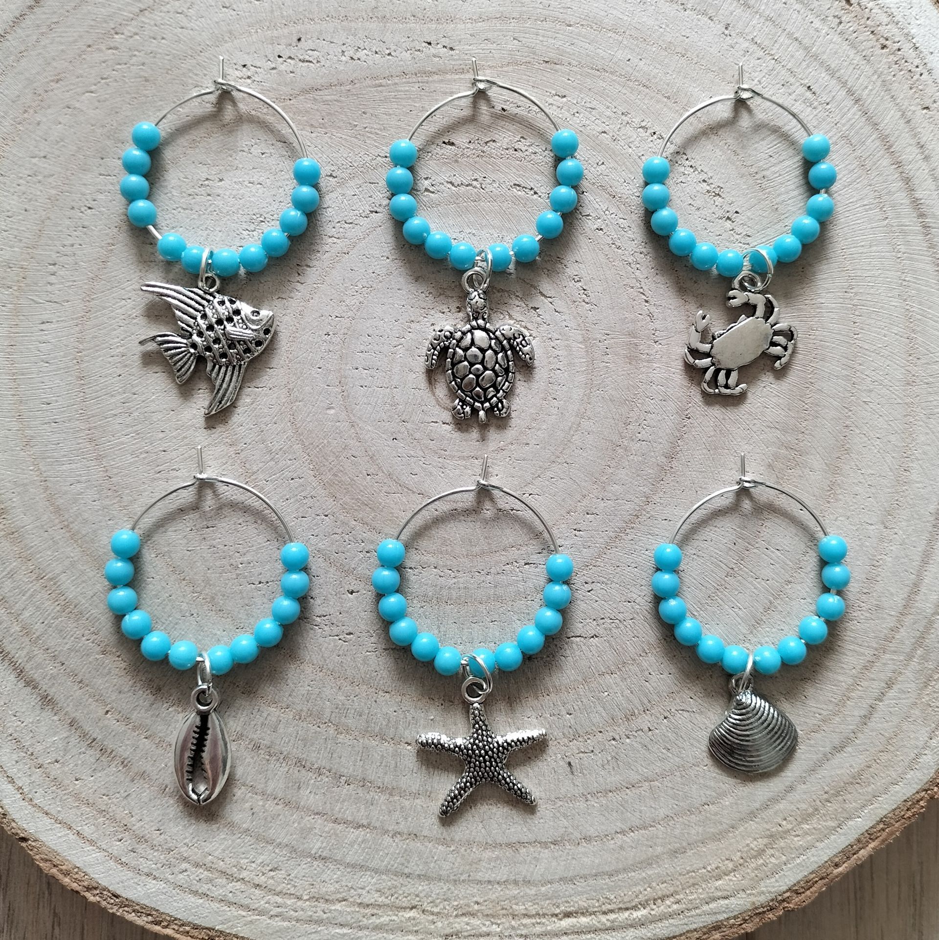 Zes turquoise wijnglasbedeltjes met zilveren hangers in maritiem thema, bevestigd aan een houten schijfje.
