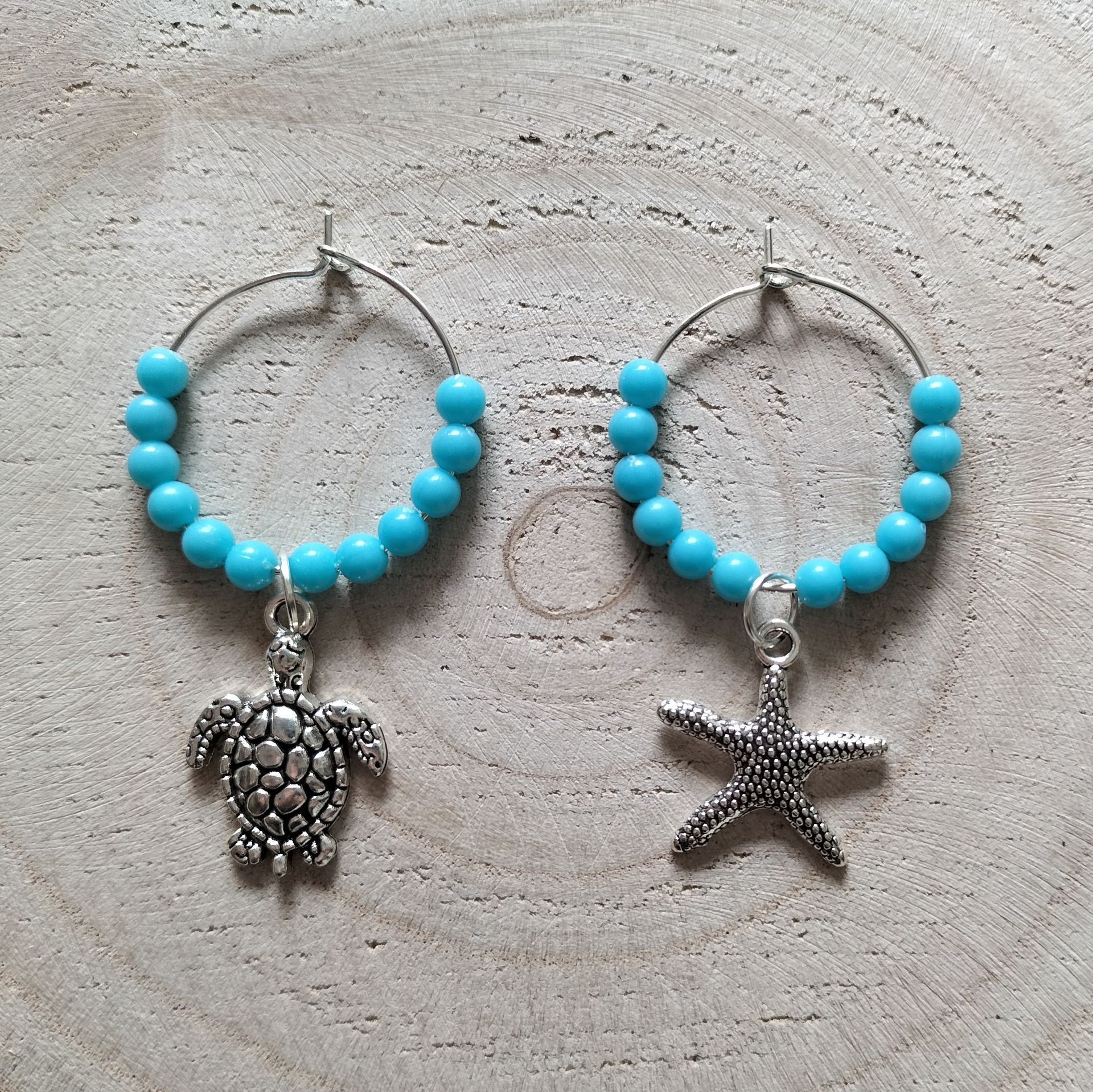 Zes turquoise wijnglasbedeltjes met zilveren hangers in maritiem thema, bevestigd aan een houten schijfje.