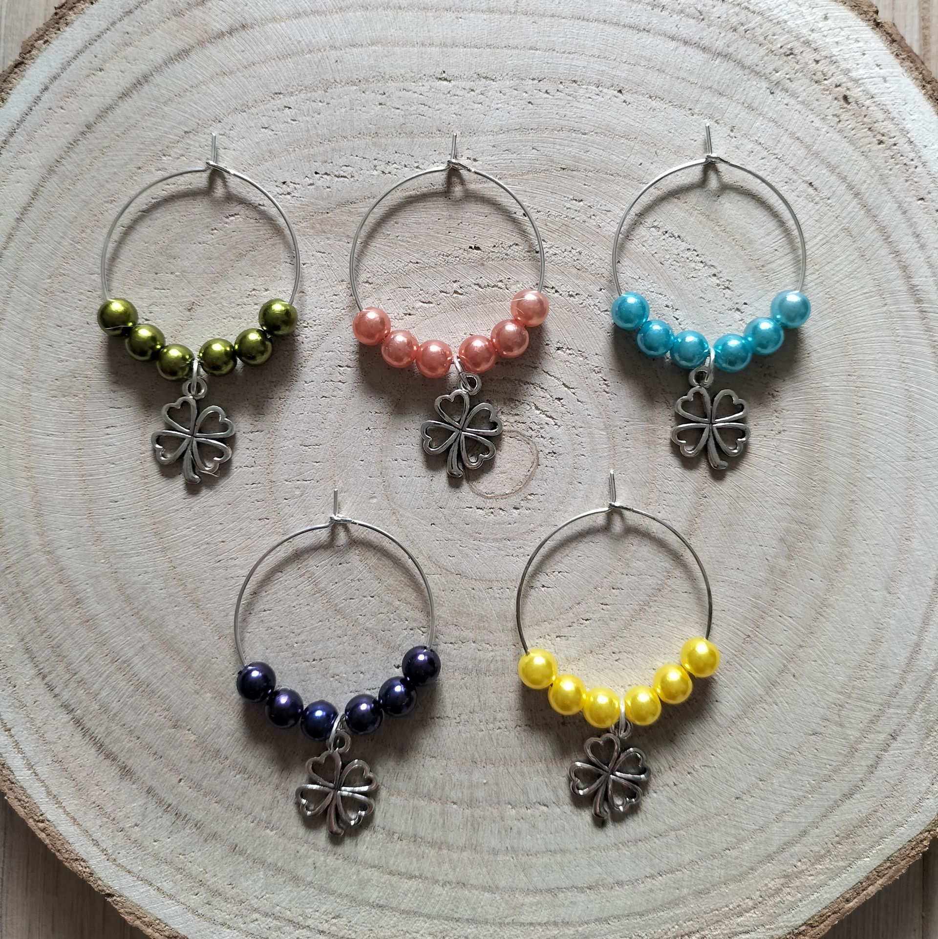 Vijf wijnhangers van kralen met klavertjes in verschillende kleuren, rustend op een houten schijfje.