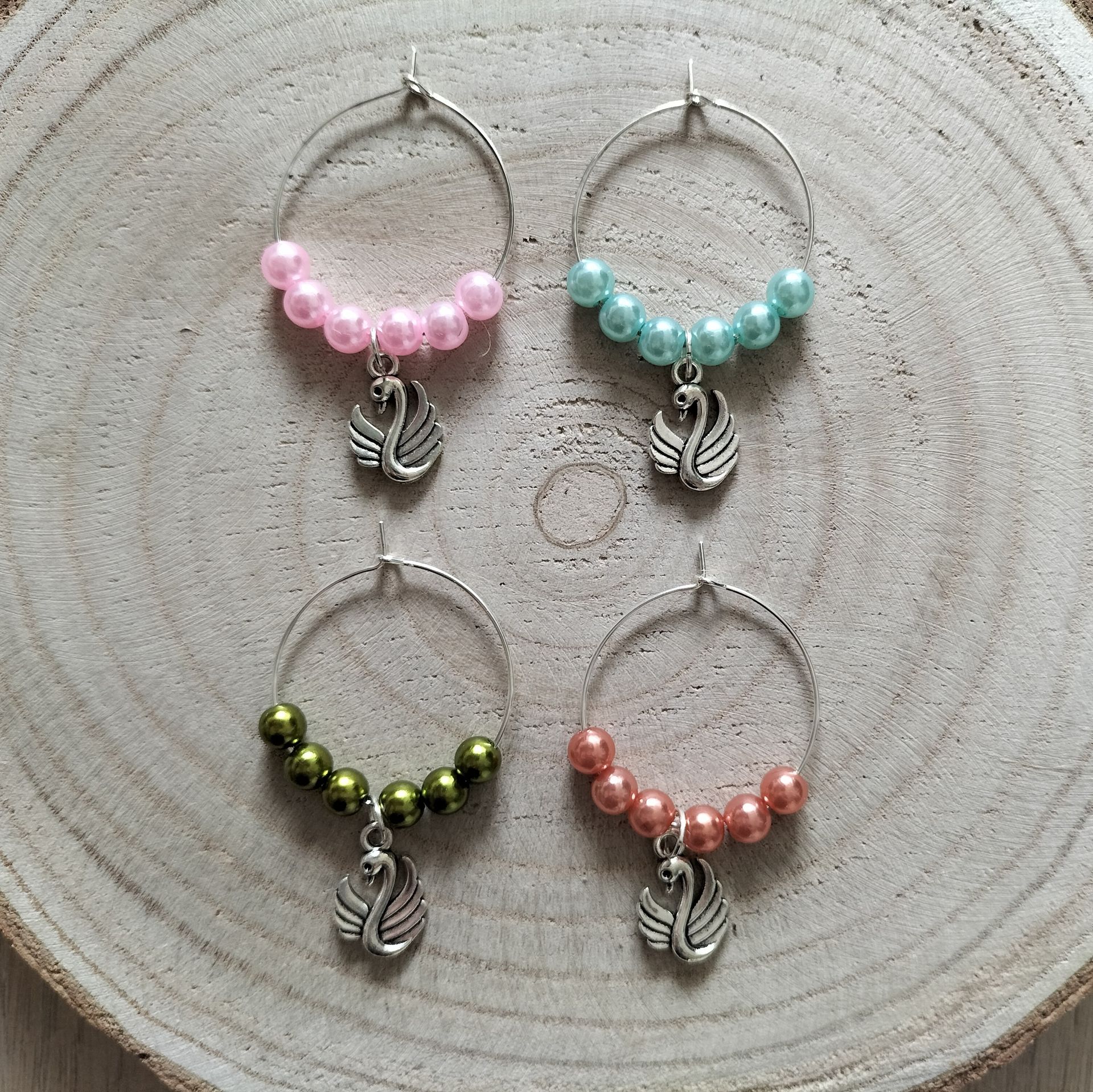 Vier wijnglashangertjes met kralen en zwanenhangertjes in roze, turquoise, groen en perzik, op een houten ondergrond.