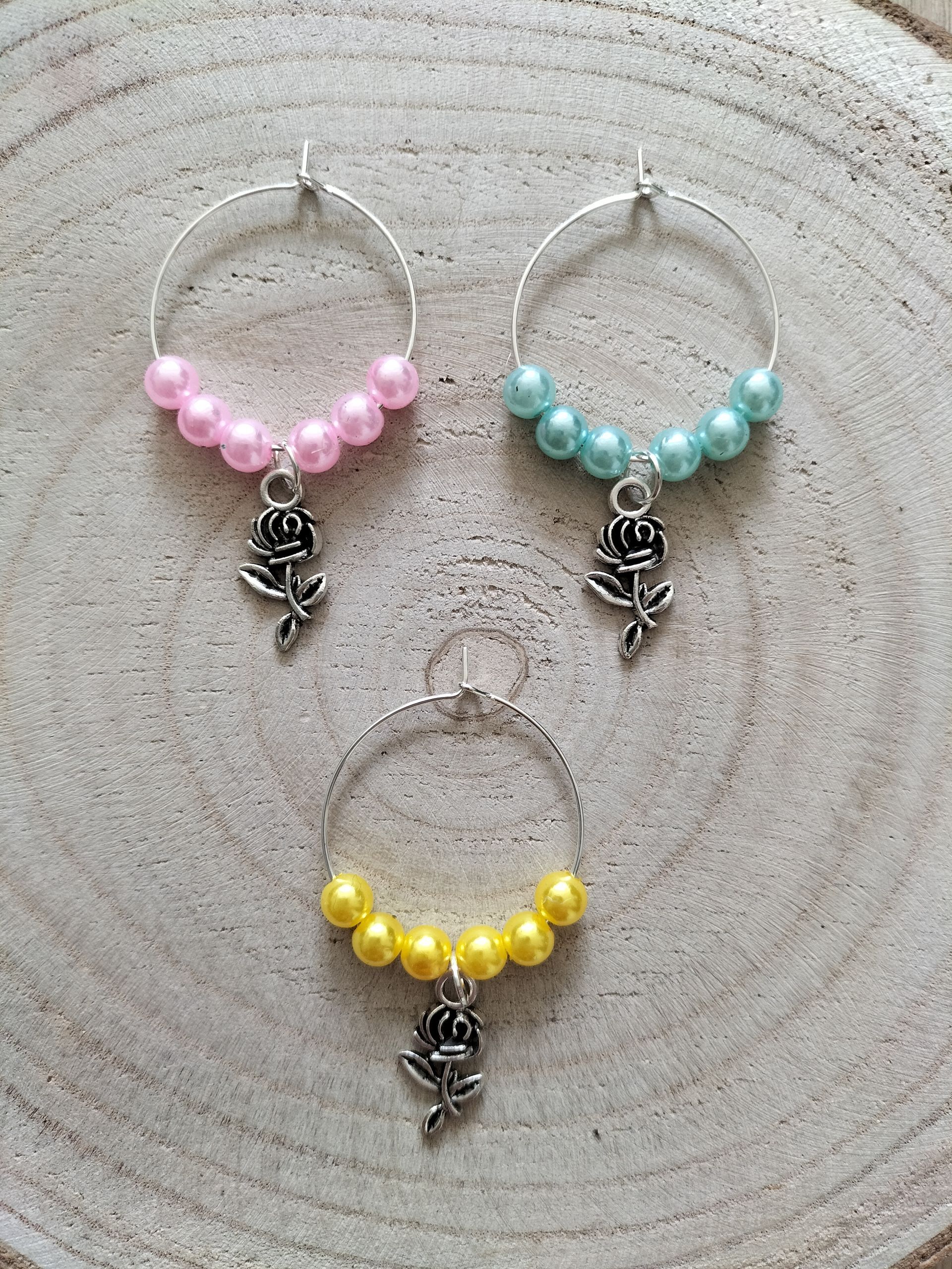 Drie ringen met wijnbedeltjes: roze, blauwe en gele kralen met zilveren rozenhangers, rustend op een houten ondergrond.