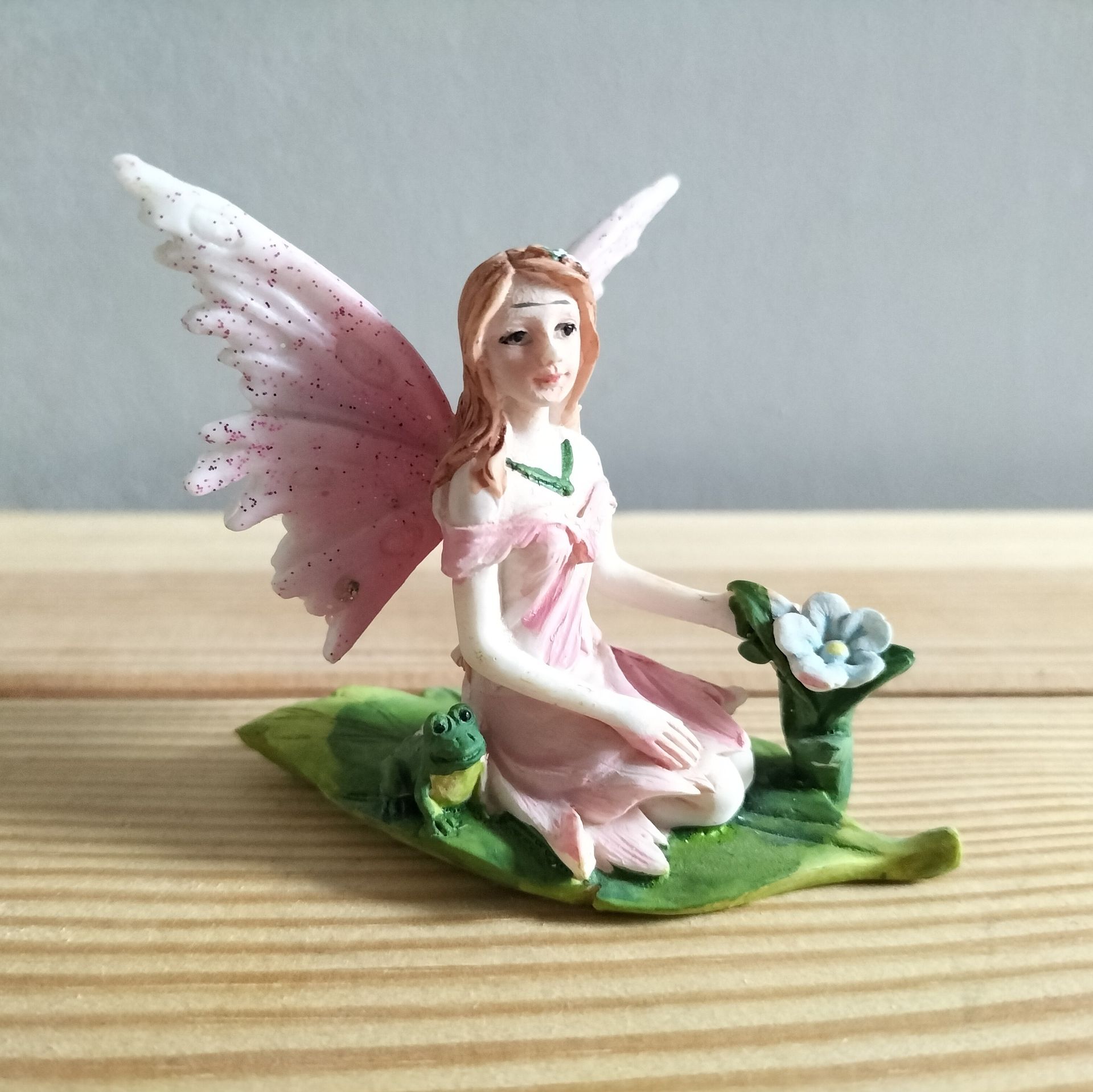 Elfje met roze vleugels, gekleed in een roze jurk, zittend op een groen blad met een kikker.