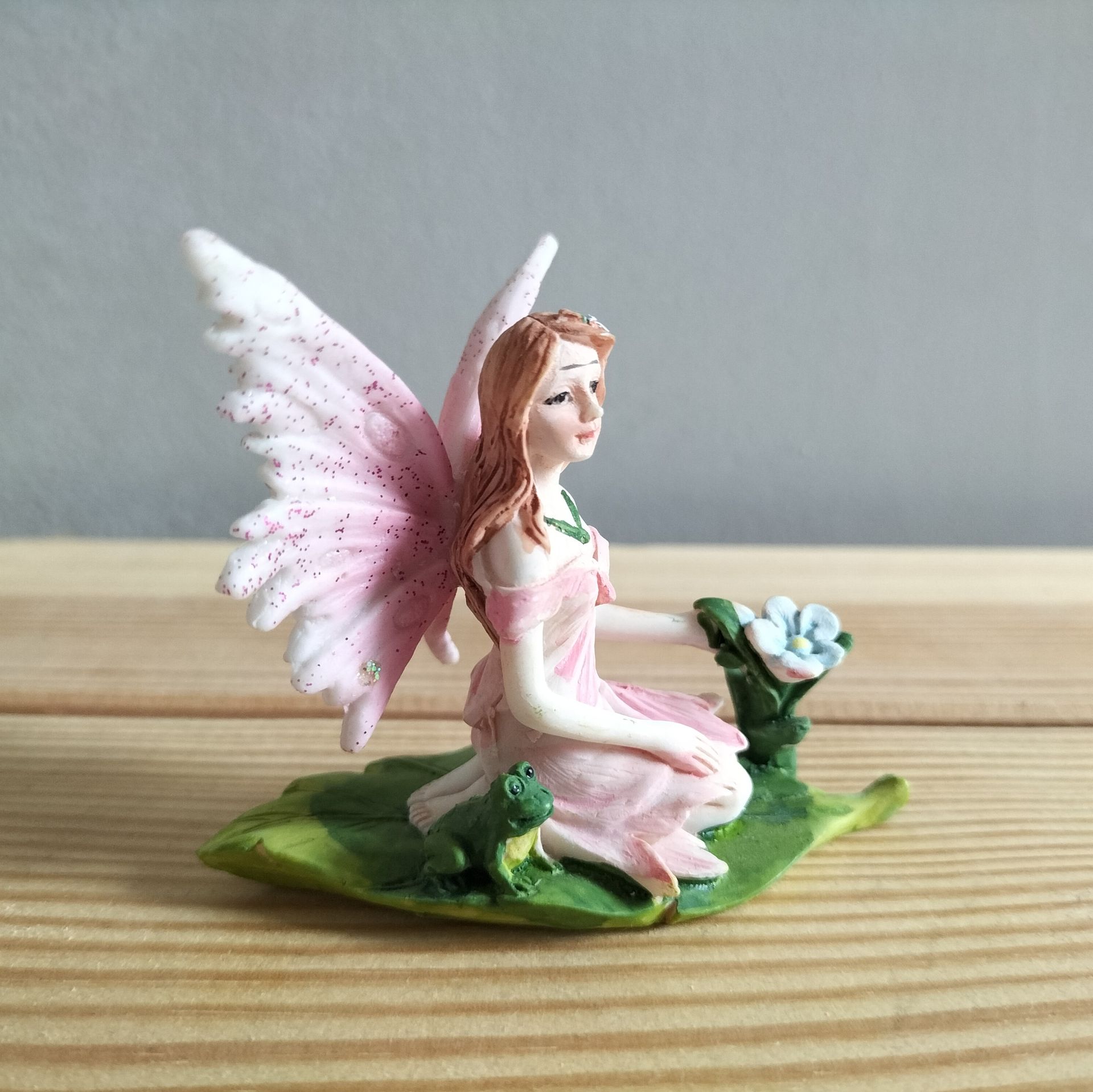 Elfje met roze vleugels, gekleed in een roze jurk, zittend op een groen blad met een kikker.