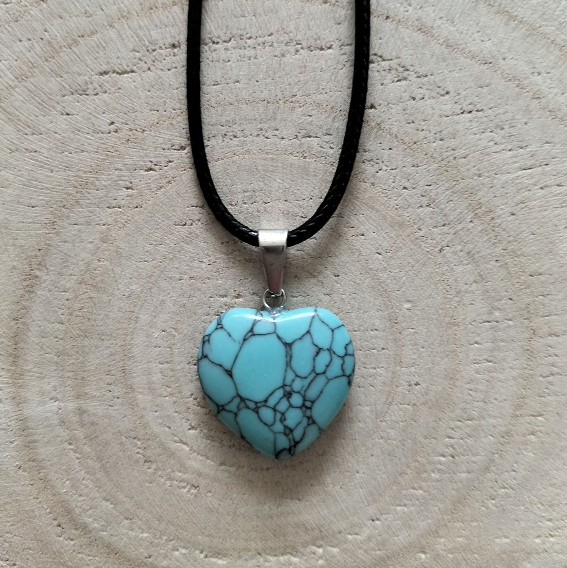 Een ketting met een turquoise hartvormige hanger aan een zwart koord, met een achtergrond van houtnerf.