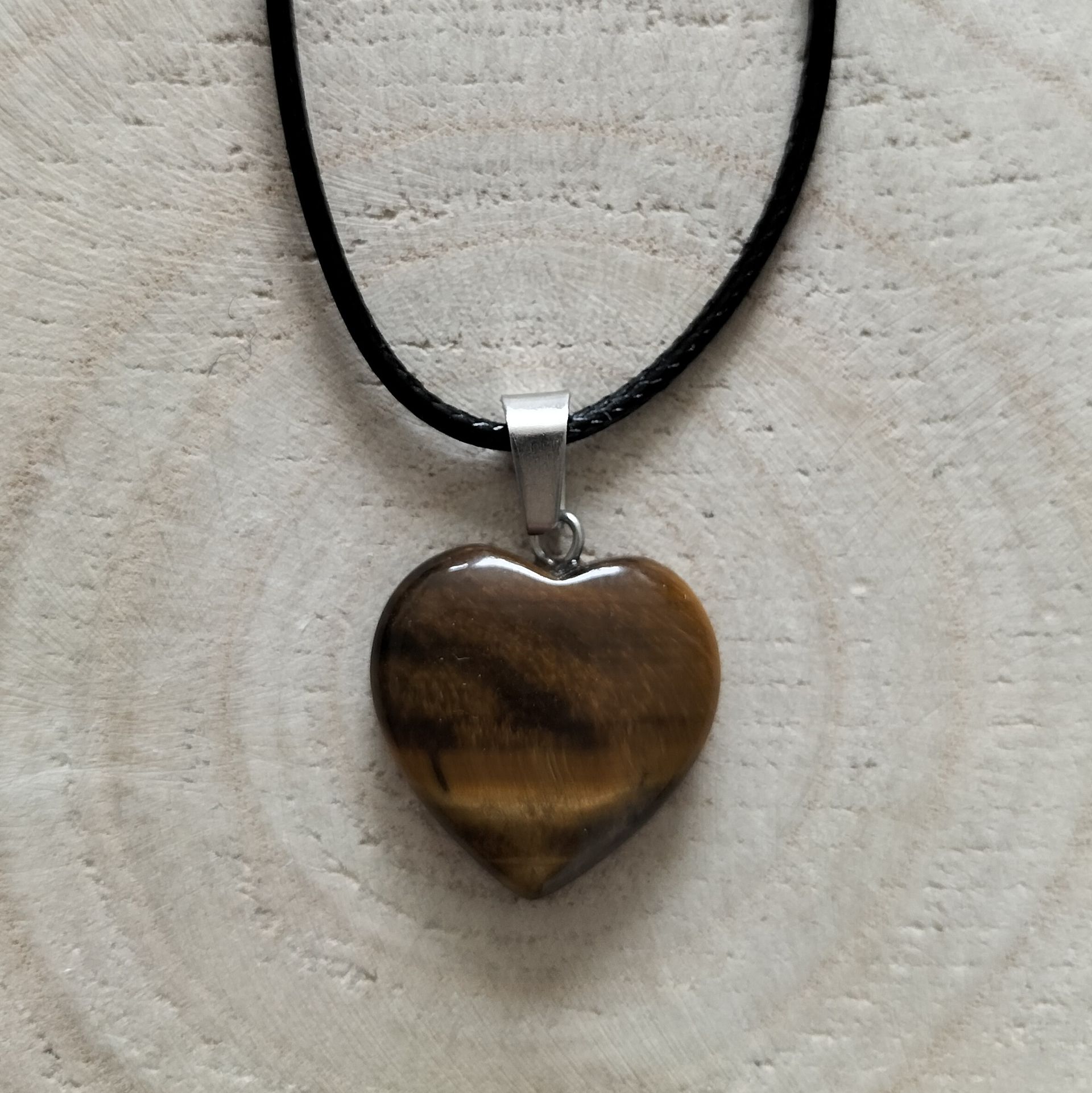 Halsketting met hartvormige hanger van tijgeroogsteen aan een zwart koord, op een houten achtergrond.