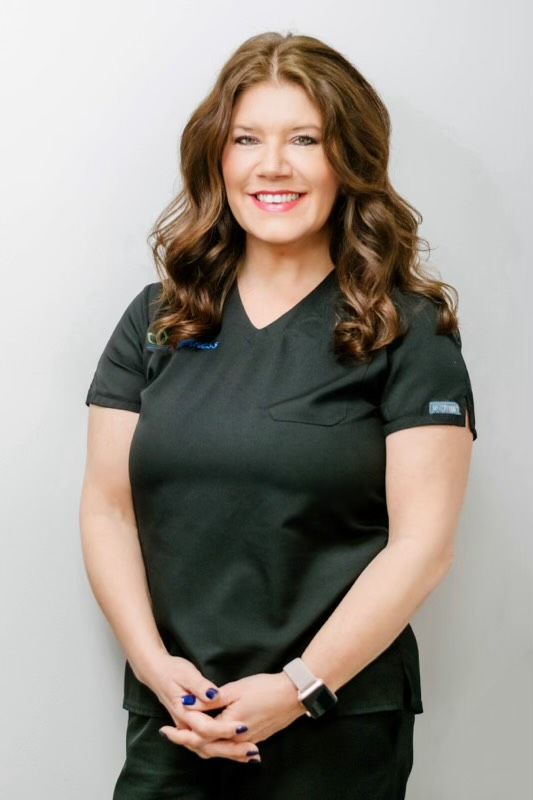 Smiling person in dark green scrubs standing against a light background, hands clasped in front