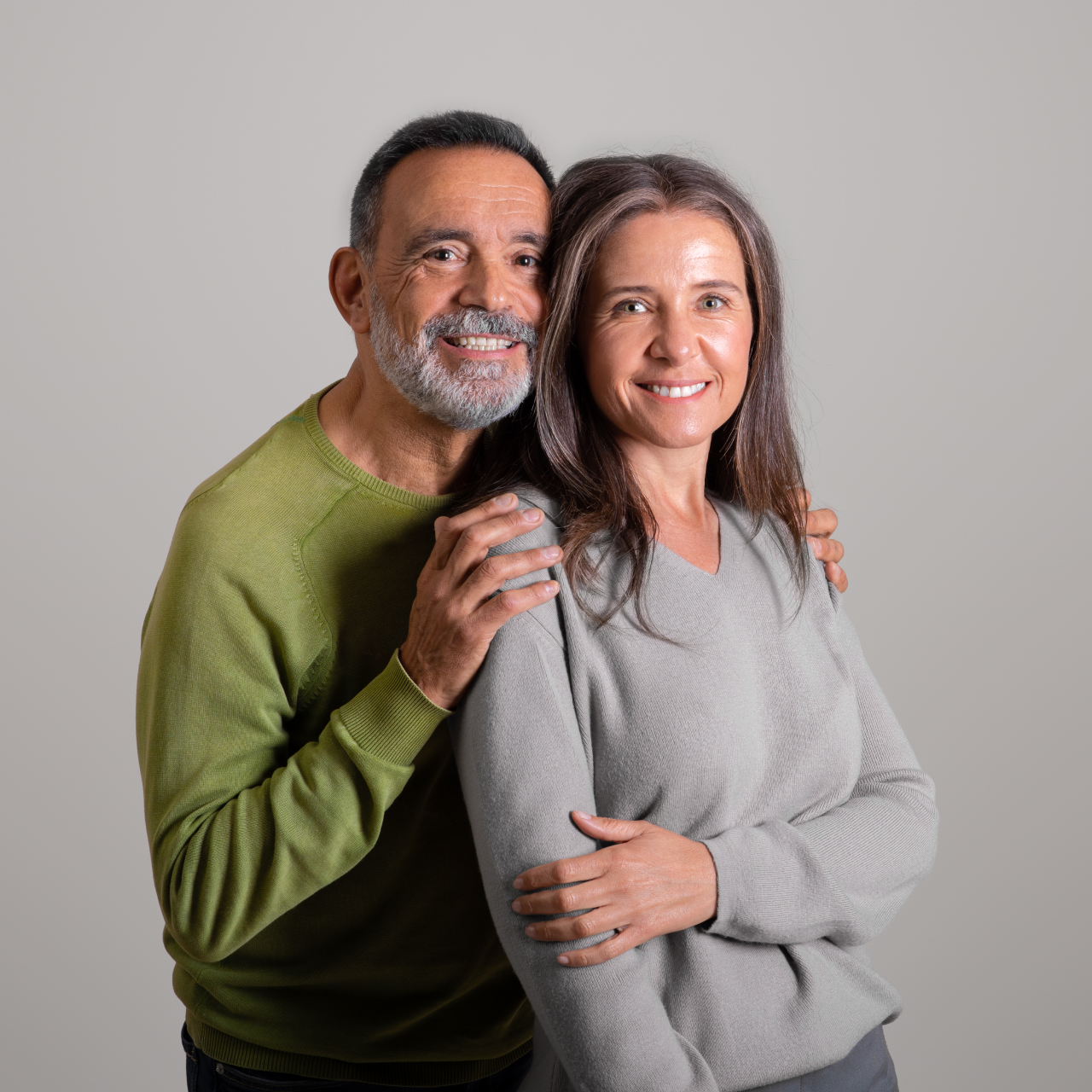 Two smiling adults pose together against a plain gray background, wearing green and gray sweaters.