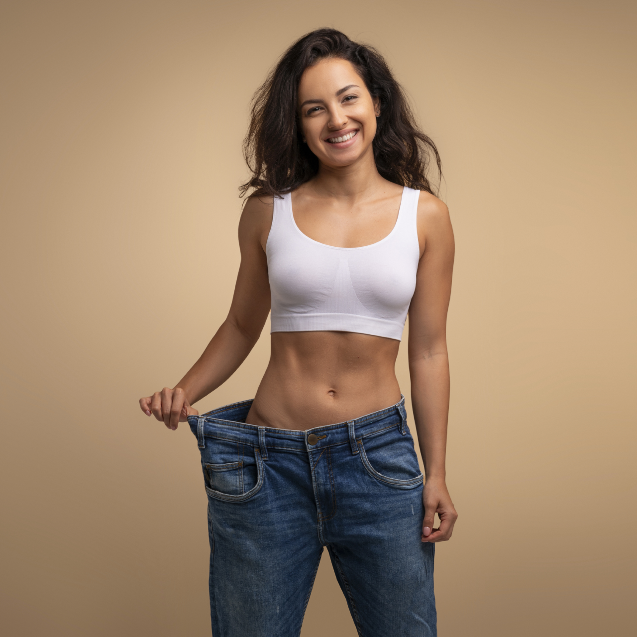 Smiling woman holding oversized jeans at the waist, wearing a white crop top against a plain tan background