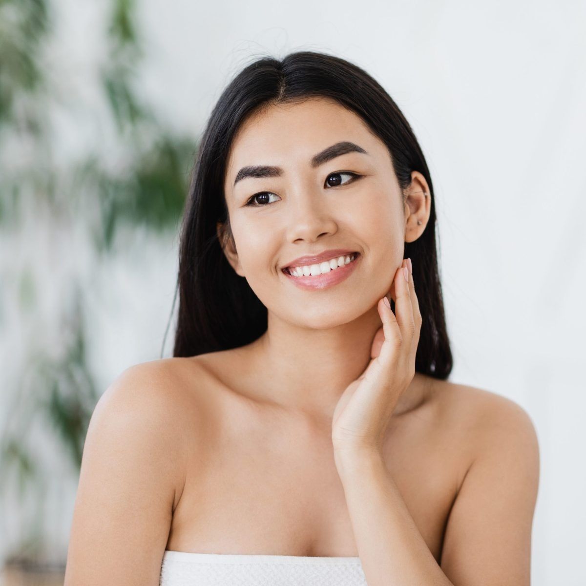 Smiling woman with long dark hair touching her cheek against a soft white background