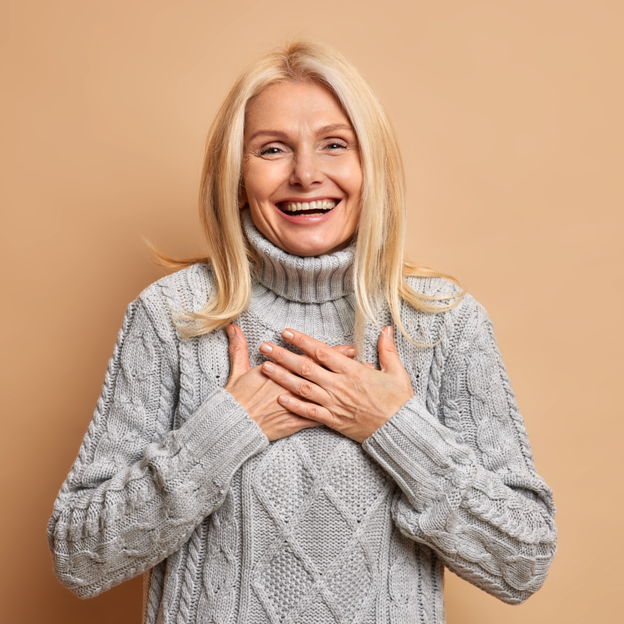 Smiling woman in a gray sweater with hands on her chest against a beige background