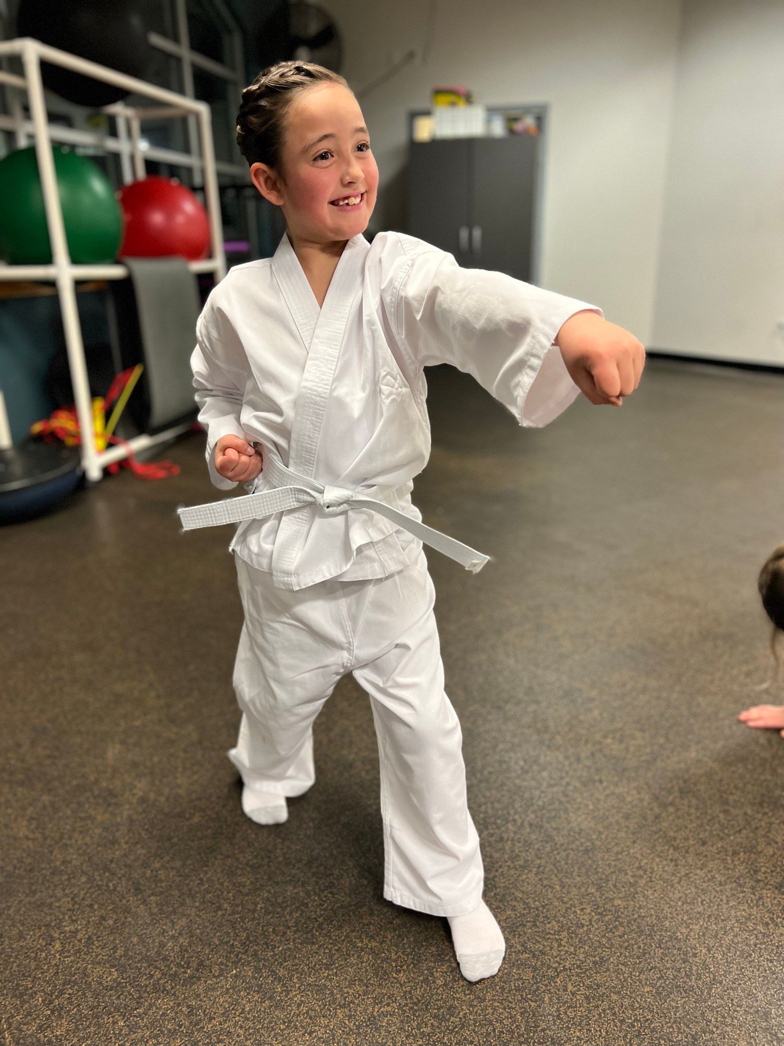 Girl in karate uniform throws a punch in a gym. She has a gray belt and a happy expression.