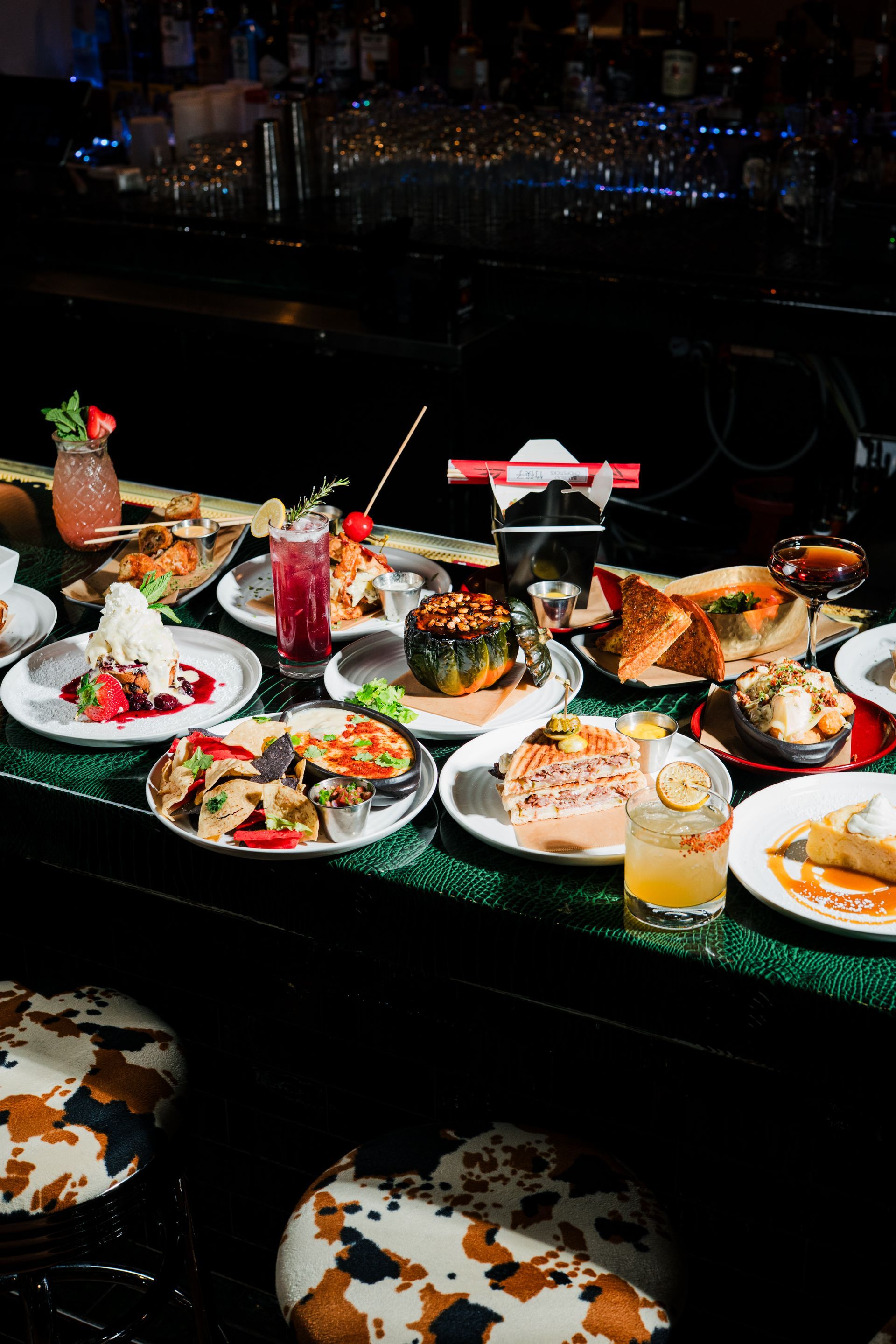 A table laden with various dishes and cocktails, lit by bar lights. Cow print bar stools in foreground.
