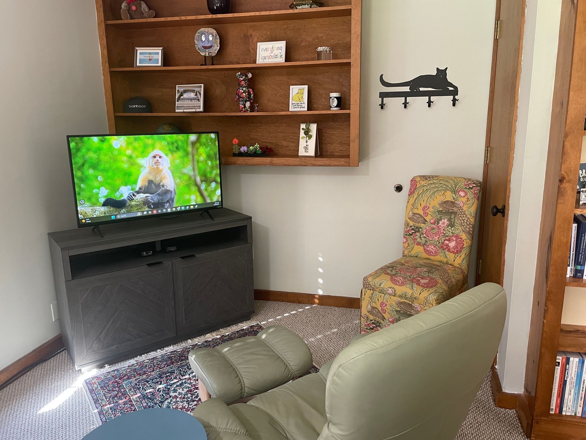 A cozy reading nook with a reclining chair facing a TV, a small bookcase, and a floral-patterned chair. A built-in bookshelf holds decorative items.