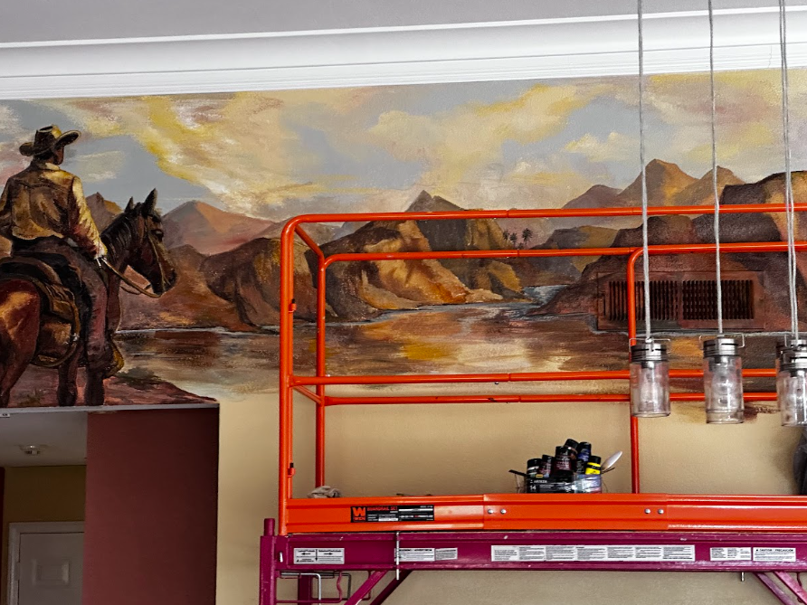 Murals of a cowboy on horseback and a desert landscape above orange scaffold and paint supplies
