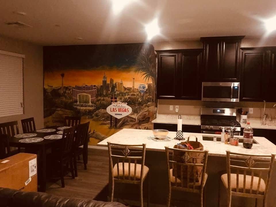 Dim kitchen with dark cabinets, granite island, bar stools, dining table, and city mural on the wall.