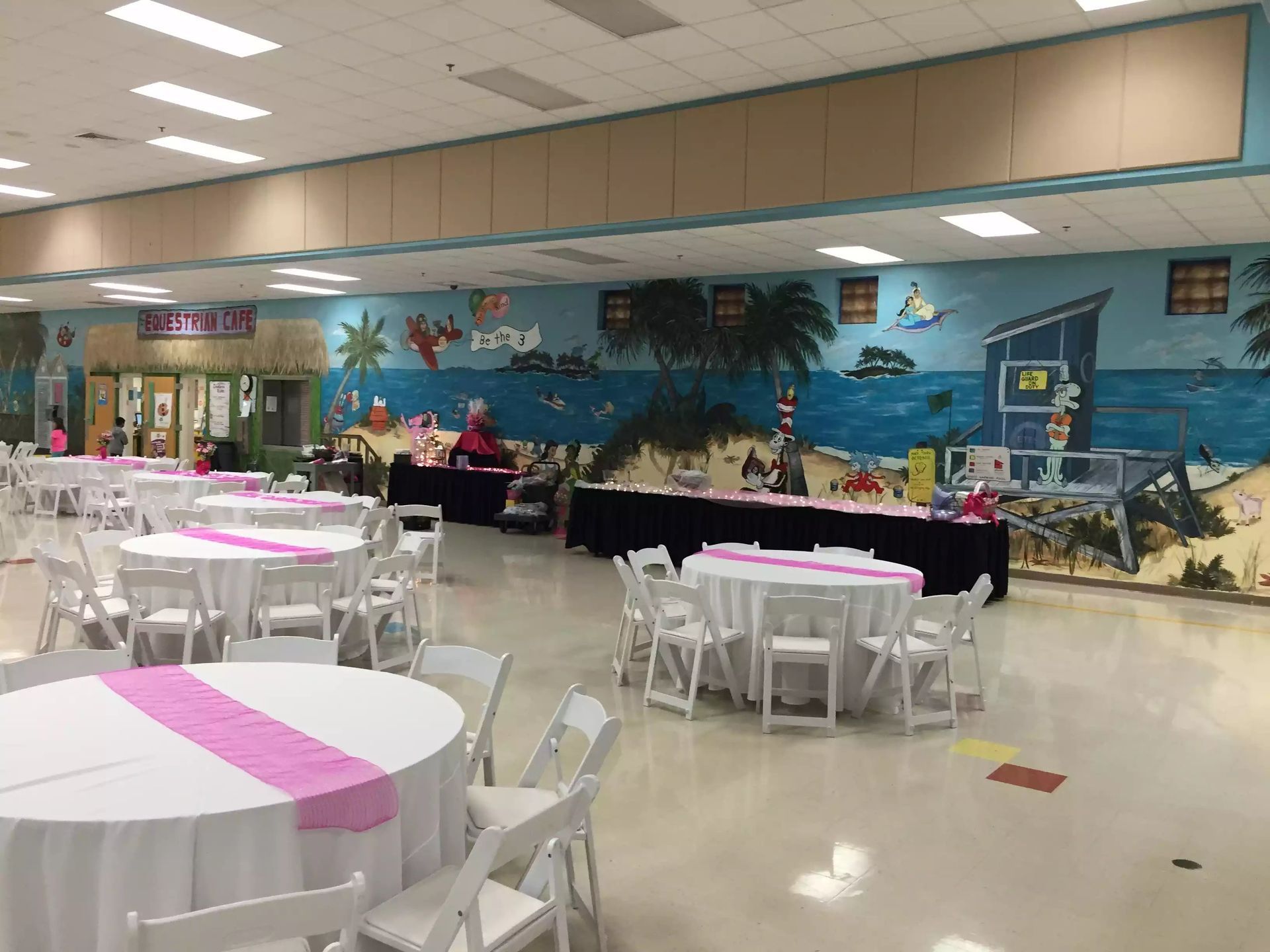 Decorated banquet hall with round tables, white chairs, and a tropical mural along the back wall.