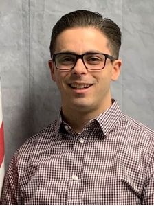 Smiling person in glasses and a checkered shirt against a gray backdrop.