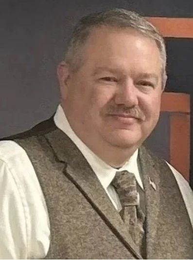 Smiling man in a white shirt and brown vest against a dark backdrop