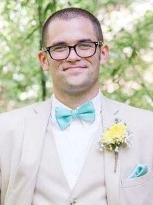 Man in a cream suit with turquoise bow tie and boutonniere, smiling outdoors.