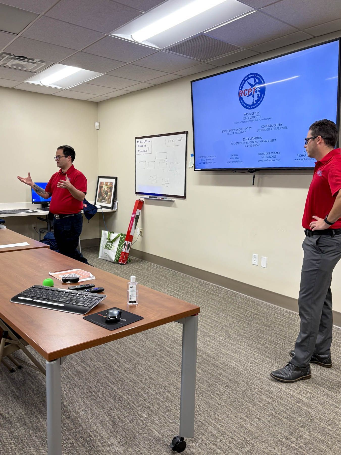 Two men in red shirts speaking in a conference room beside a large presentation screen.