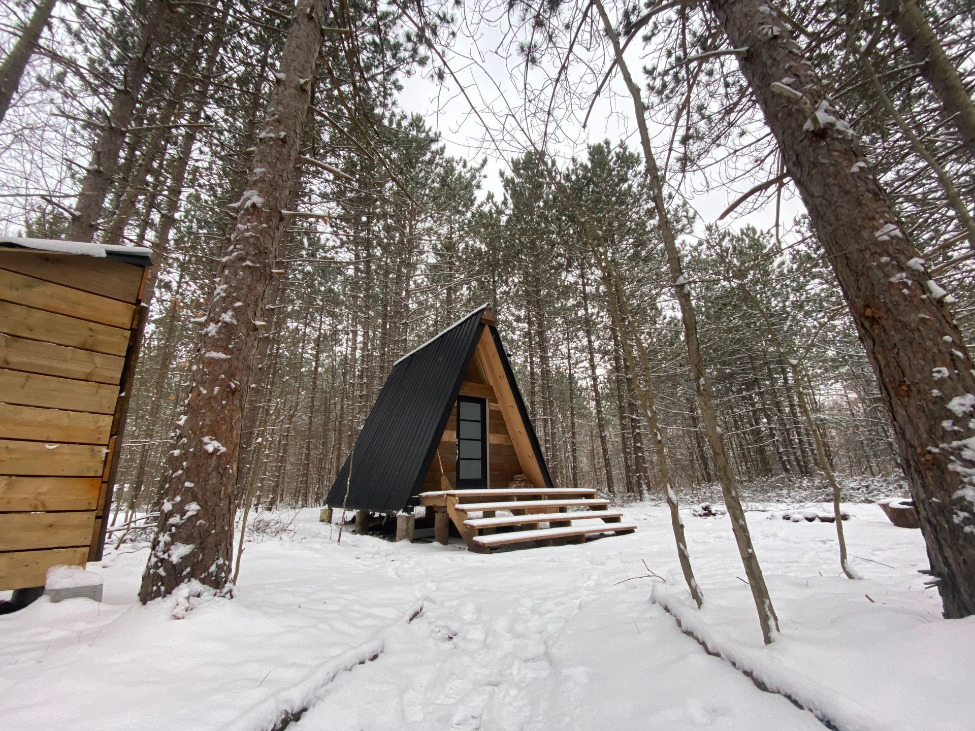 A-frame cabin in snowy woods; black roof, wooden porch, surrounding tall pine trees.