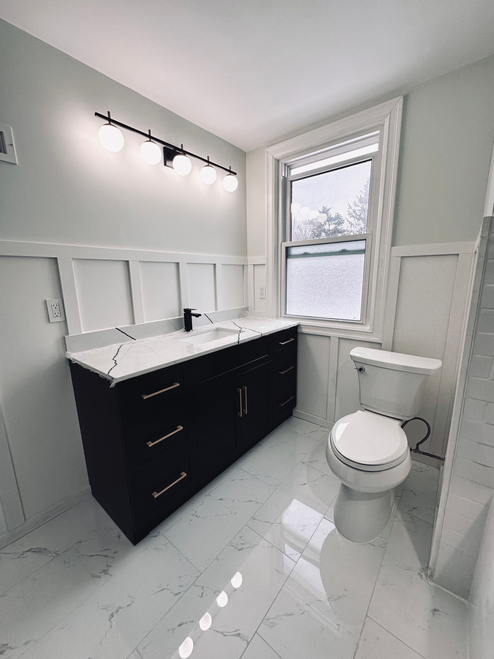 Bathroom with dark vanity, white countertop, and modern light fixture. White tile floor and wall paneling.