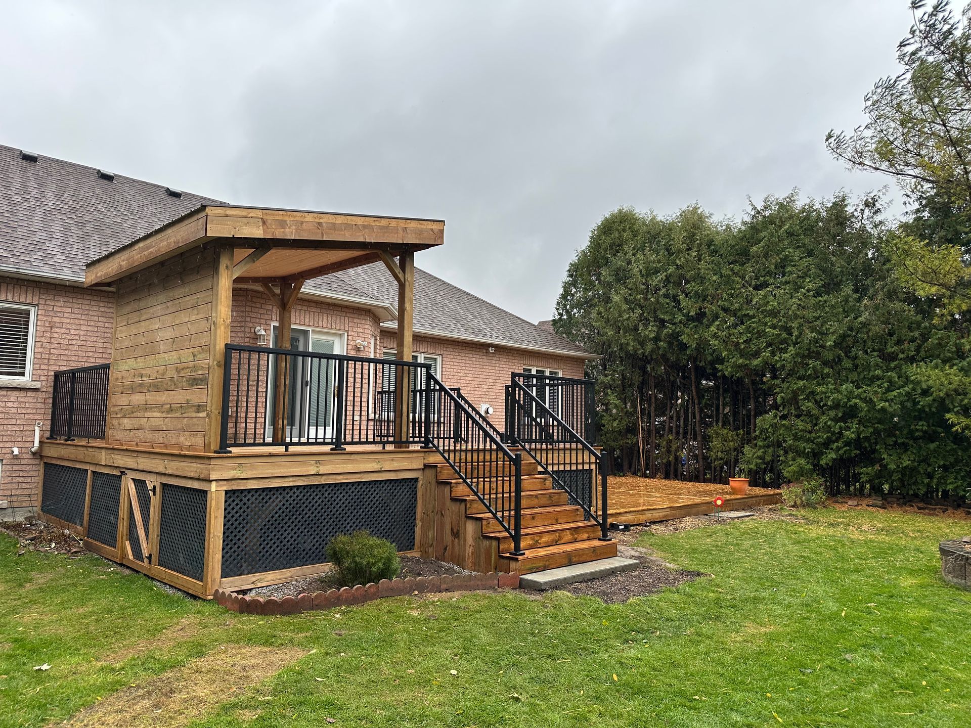 Wooden deck with stairs, a covered section, and black railings next to a brick house on a grassy lawn.