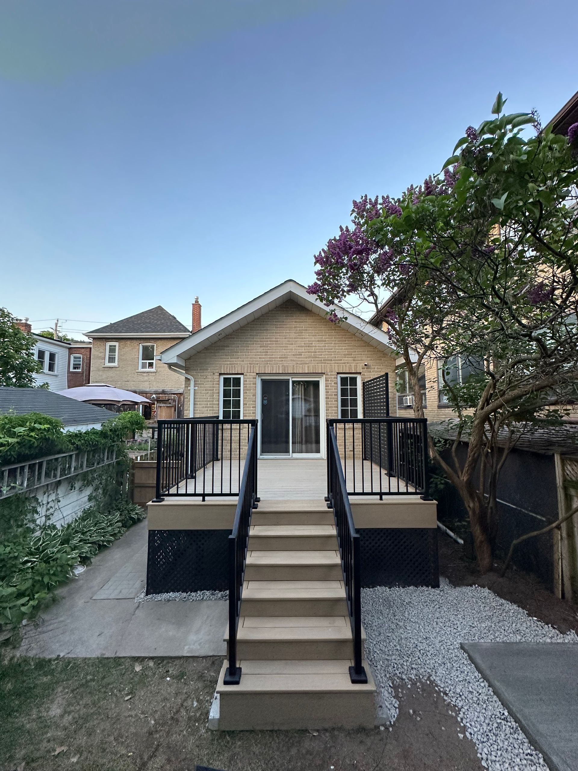 Backyard deck and house with steps. Black railing, beige siding, white rock border.