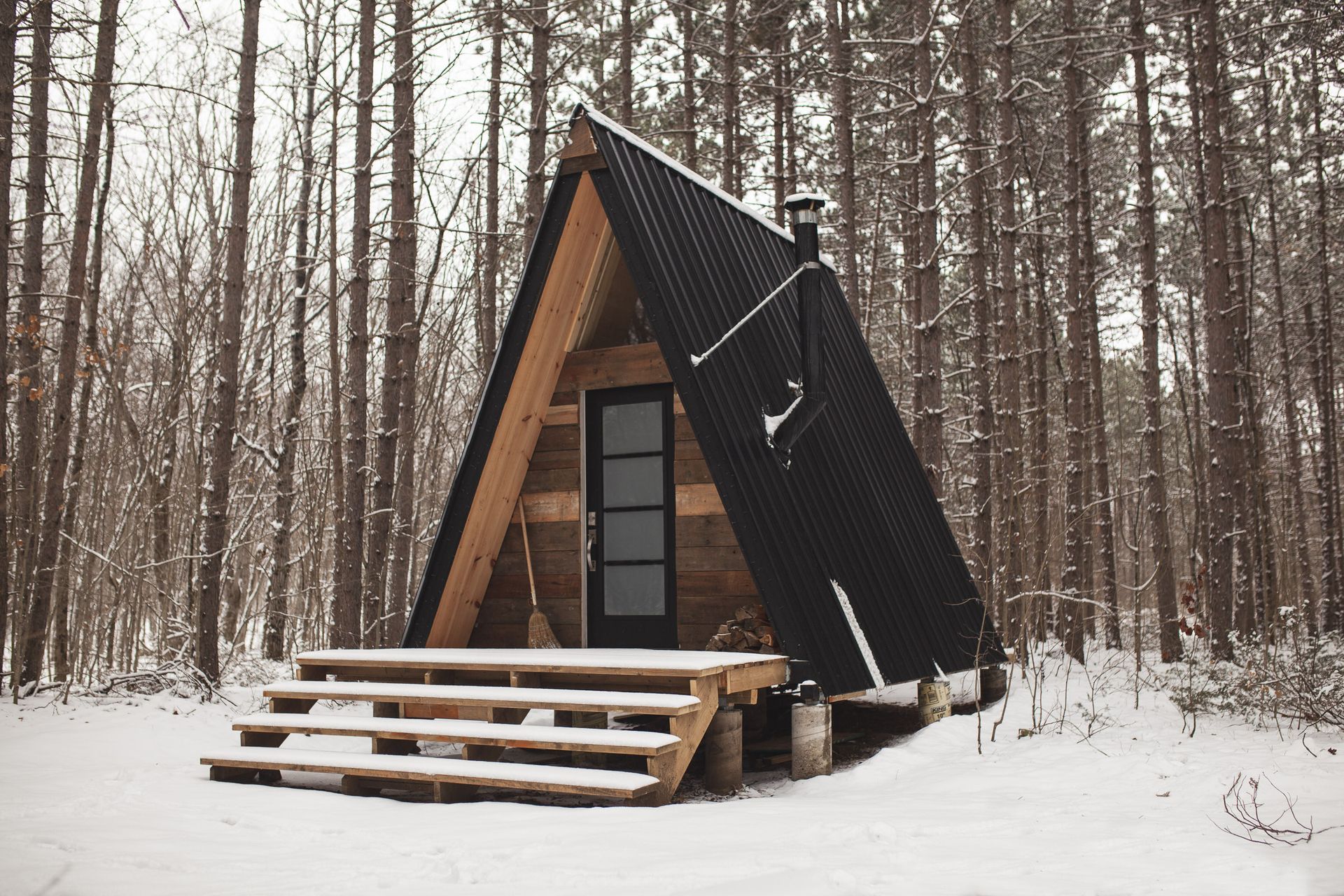 A-frame cabin with black roof in a snowy forest. Wooden steps lead to a dark door.