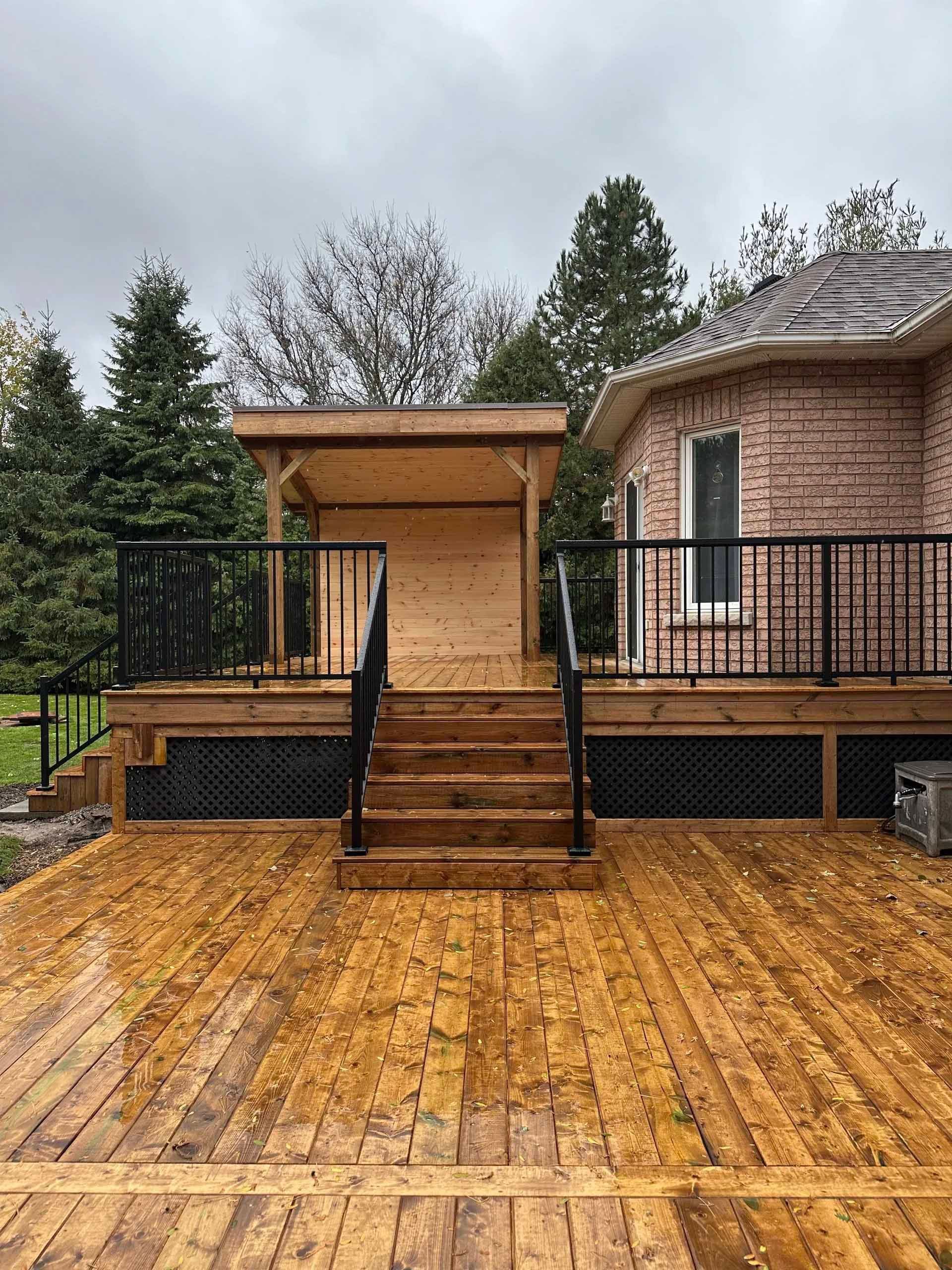 Wooden deck with black railings and steps leading to a covered structure attached to a brick house.