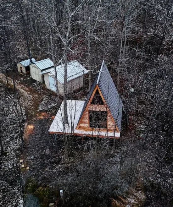 Wooden A-frame cabin and outbuildings in a snowy forest setting.
