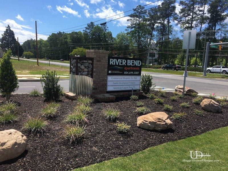 Sign for River Bend apartments with landscaping; trees, brown mulch, and rocks in a sunny setting.