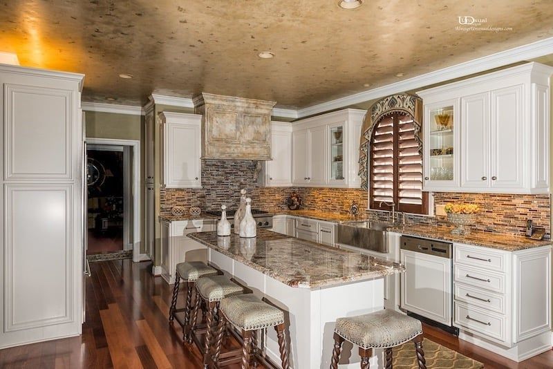 Luxurious white kitchen with island, wood floor, granite countertop, gold ceiling, and shuttered window.