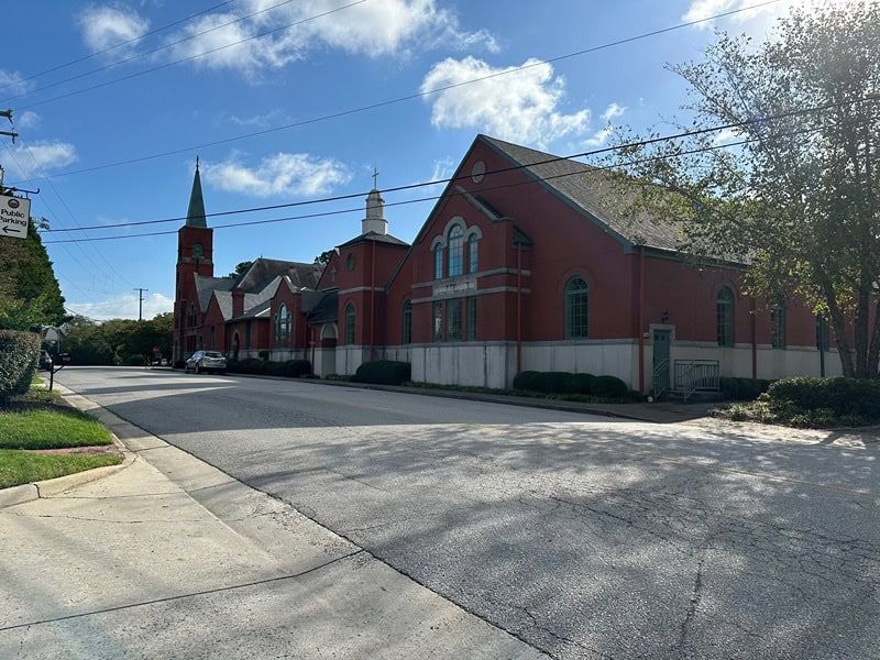 Red brick church building with steeple, on a sunny day. Street and sidewalk in foreground.