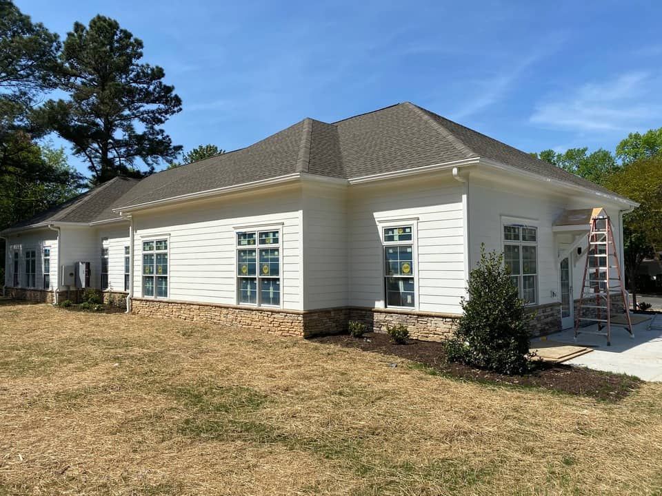 White building with a brown roof and stone base; windows, ladder, and landscaping.