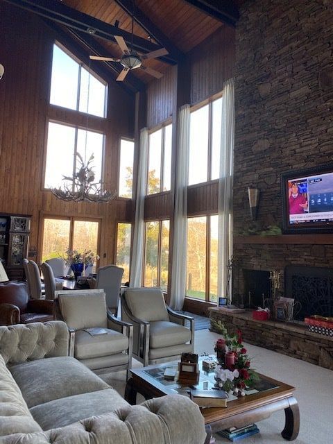 Living room with high ceilings, large windows, and a stone fireplace. Furnishings include a sofa, chairs, and a dining table.