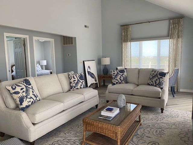 Living room with two cream-colored sofas, blue pillows, wicker coffee table, and tall windows with sheer curtains.