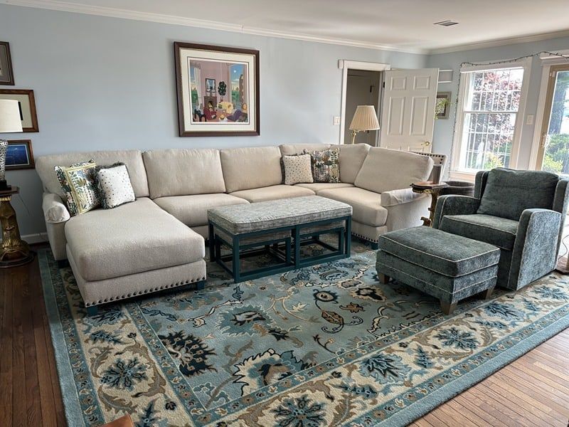 Living room with light blue walls, beige sectional, patterned blue rug, teal armchair and ottoman, and artwork.