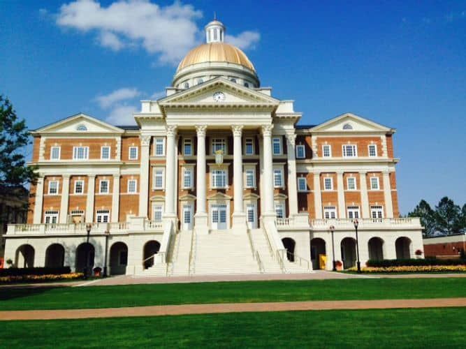 Grand brick building with a gold dome, white columns, and a large staircase on a green lawn under a blue sky.