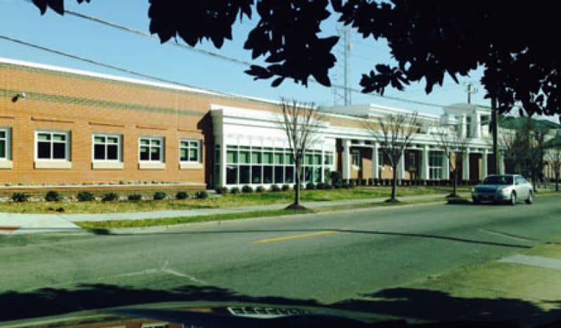 Brick building with large windows and a white-columned entrance; car drives on street in front.