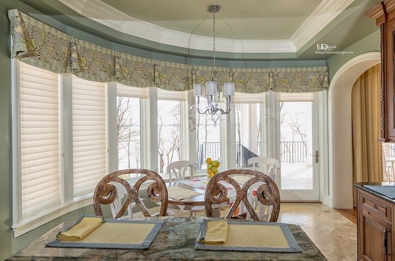 Dining area with curved windows, floral valance, chandelier, and a table set for a meal.