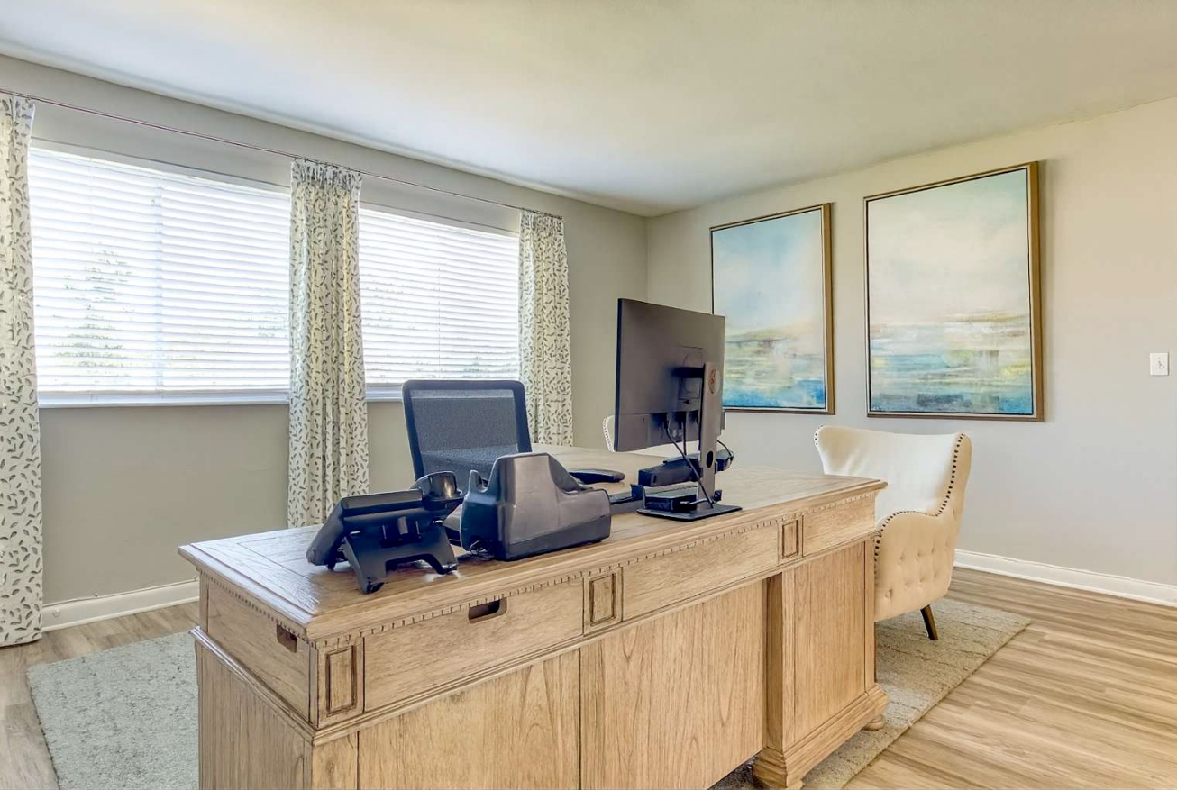 Home office with desk, computer, artwork, and chair near windows with blinds.