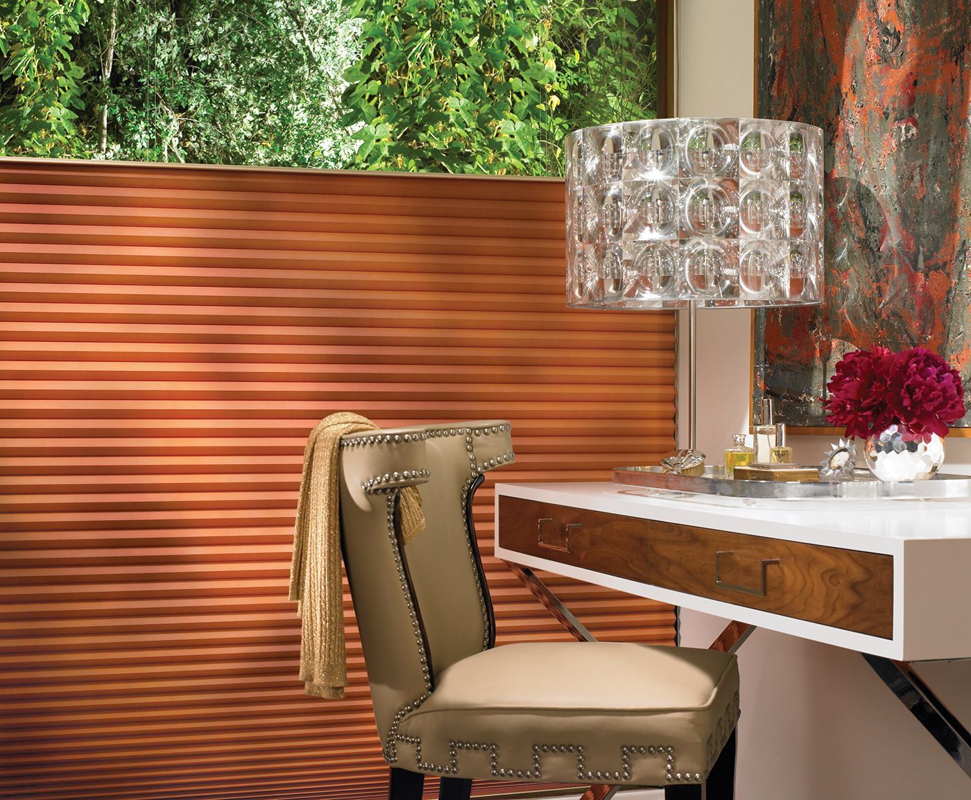 Desk with chair, lamp, and floral arrangement near copper-colored window shades and artwork; green foliage visible outside.