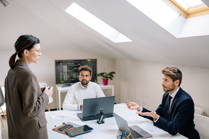 Drie collega's in een lichte kantoorruimte, die rond een bureau met een laptop en monitor documenten bespreken.