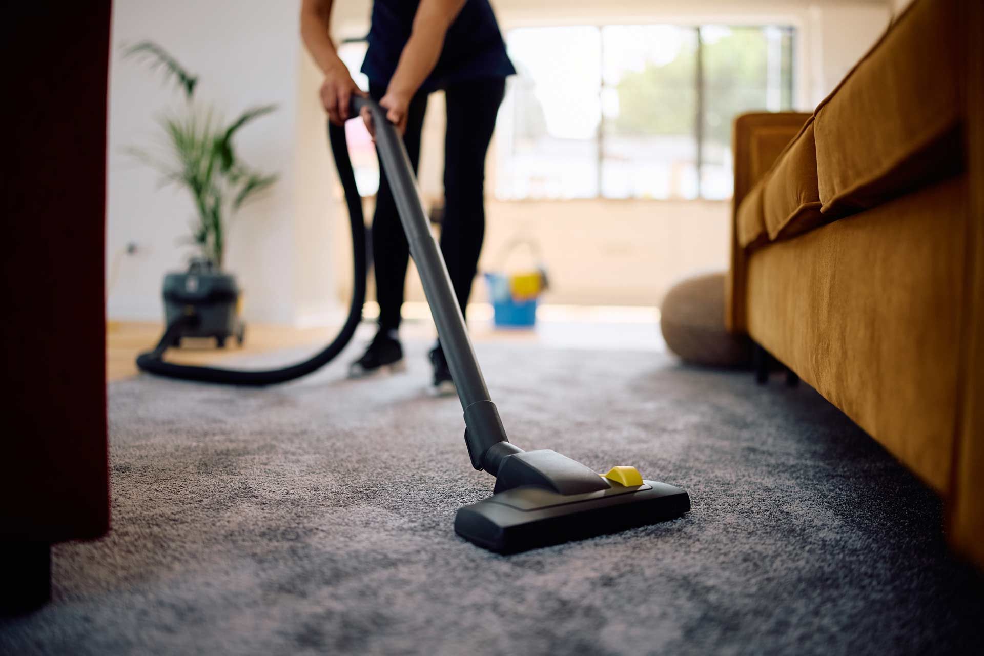 Person vacuuming a carpeted room; the vacuum cleaner is in use near a sofa.