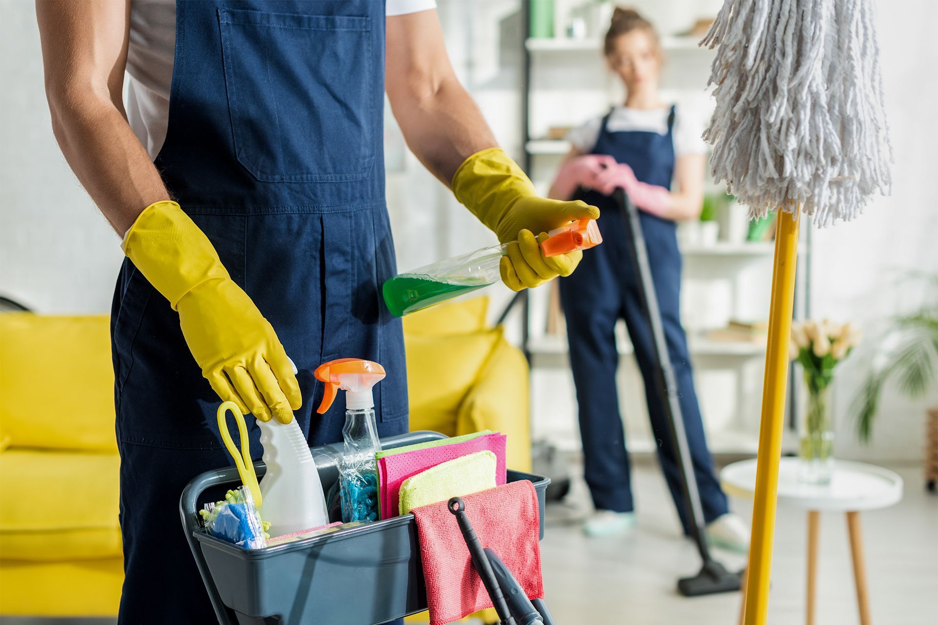 Two people cleaning a room with supplies: one holding cleaning products, the other mopping.