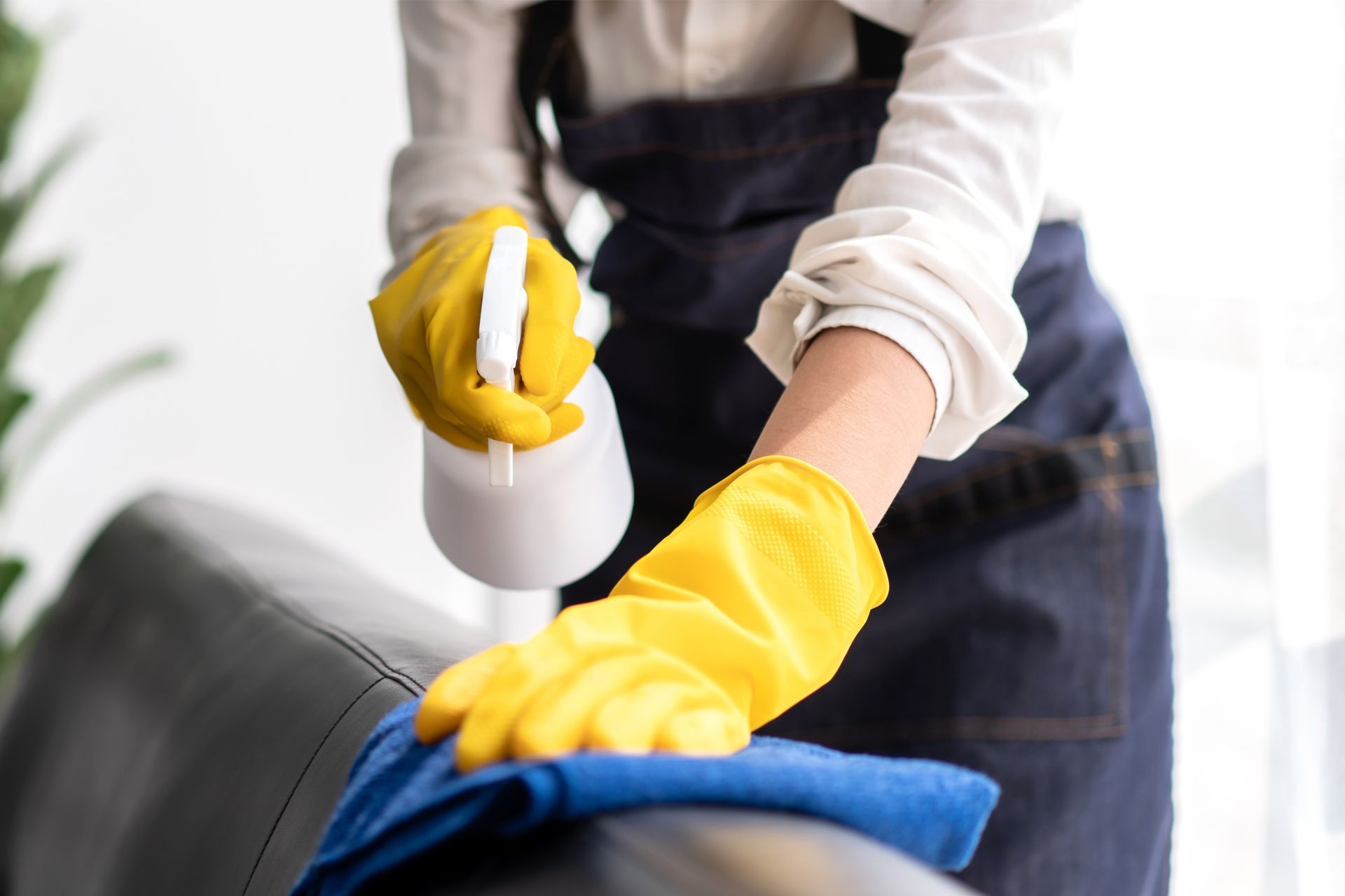 Person wearing yellow gloves and an apron sprays and wipes a black leather chair with a blue cloth.